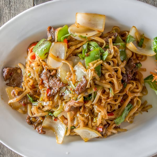 Close-up of beef pad see ew with wide noodles, green bell peppers, and onions on a white plate.